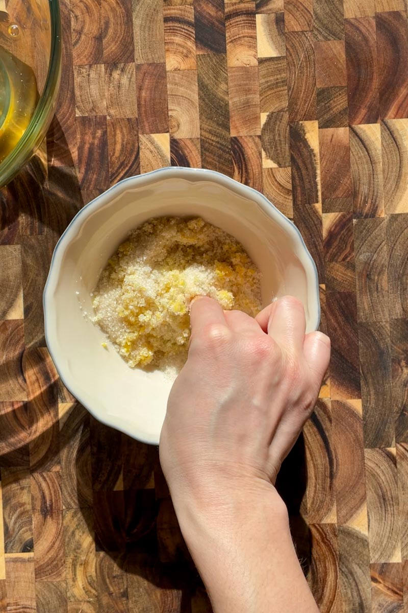 Hands rubbing lemon zest into cane sugar in a bowl to release essential oils for sourdough lemon cake