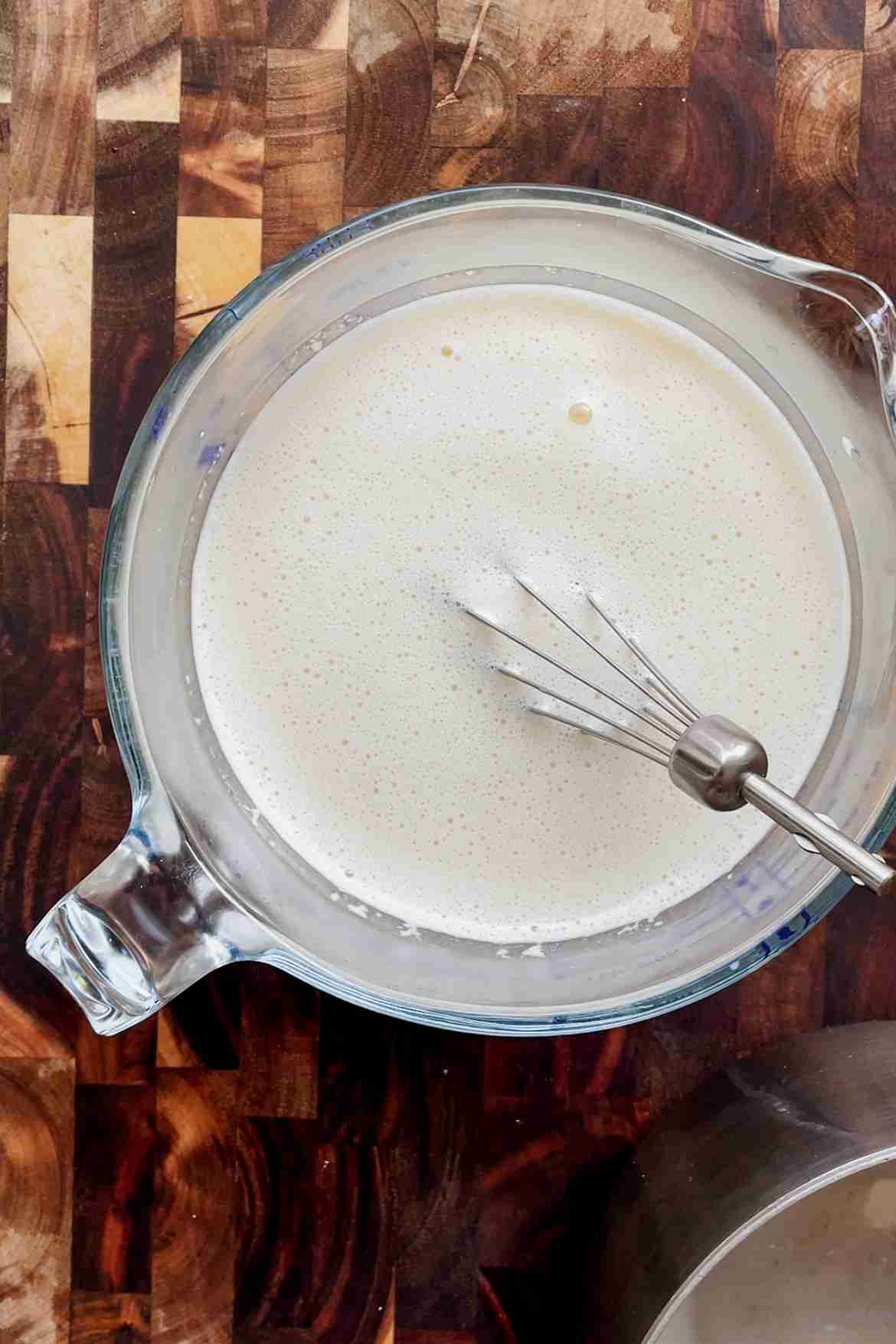Warm cream and milk being slowly poured into egg yolks while whisking to temper.