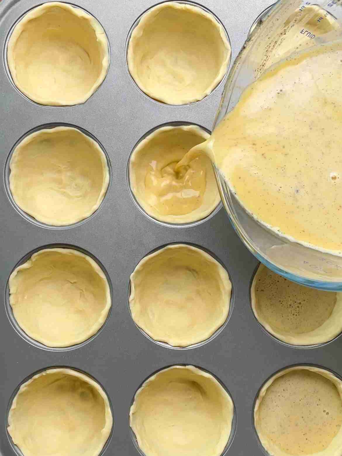 Custard mixture being poured into puff pastry shells in a muffin tin ready to bake